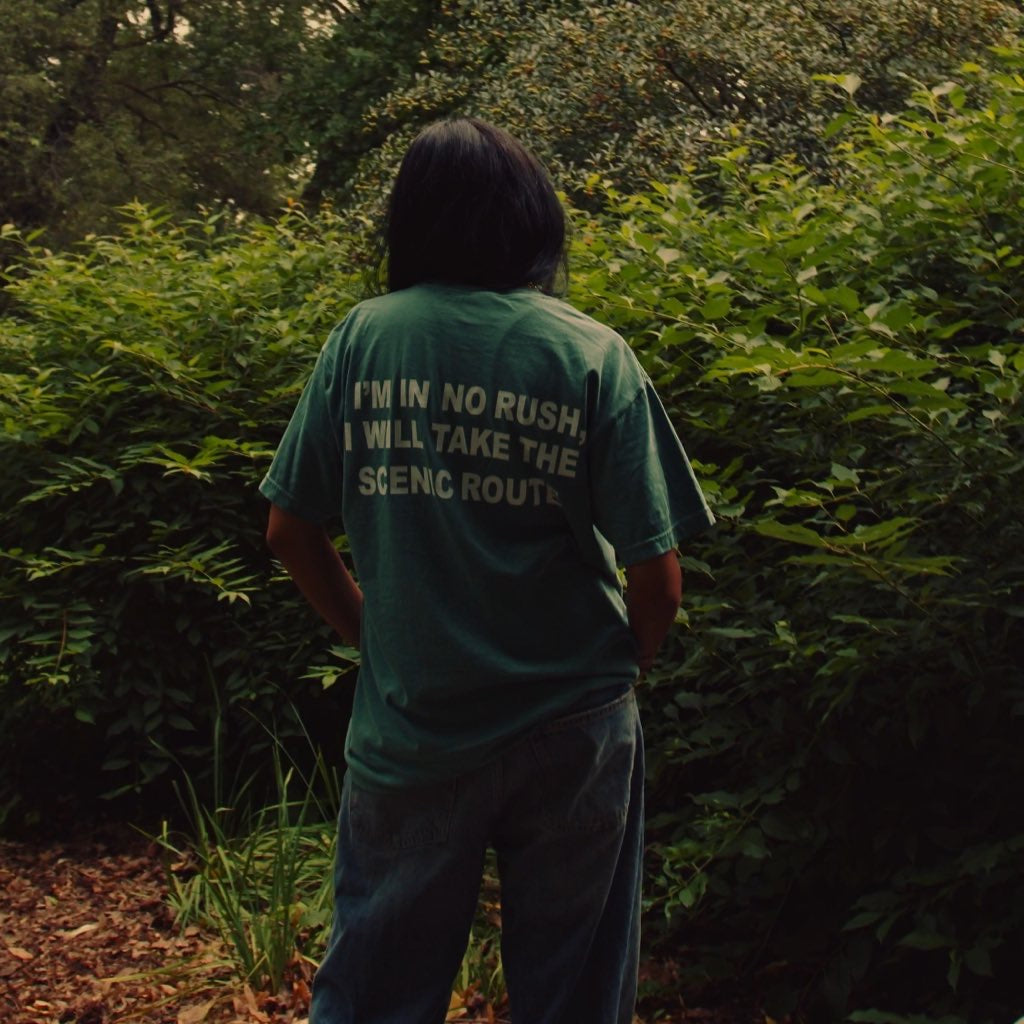 Someone standing still facing a wall of green hedge, wearing the Entry 001 tee, hands behind their back.