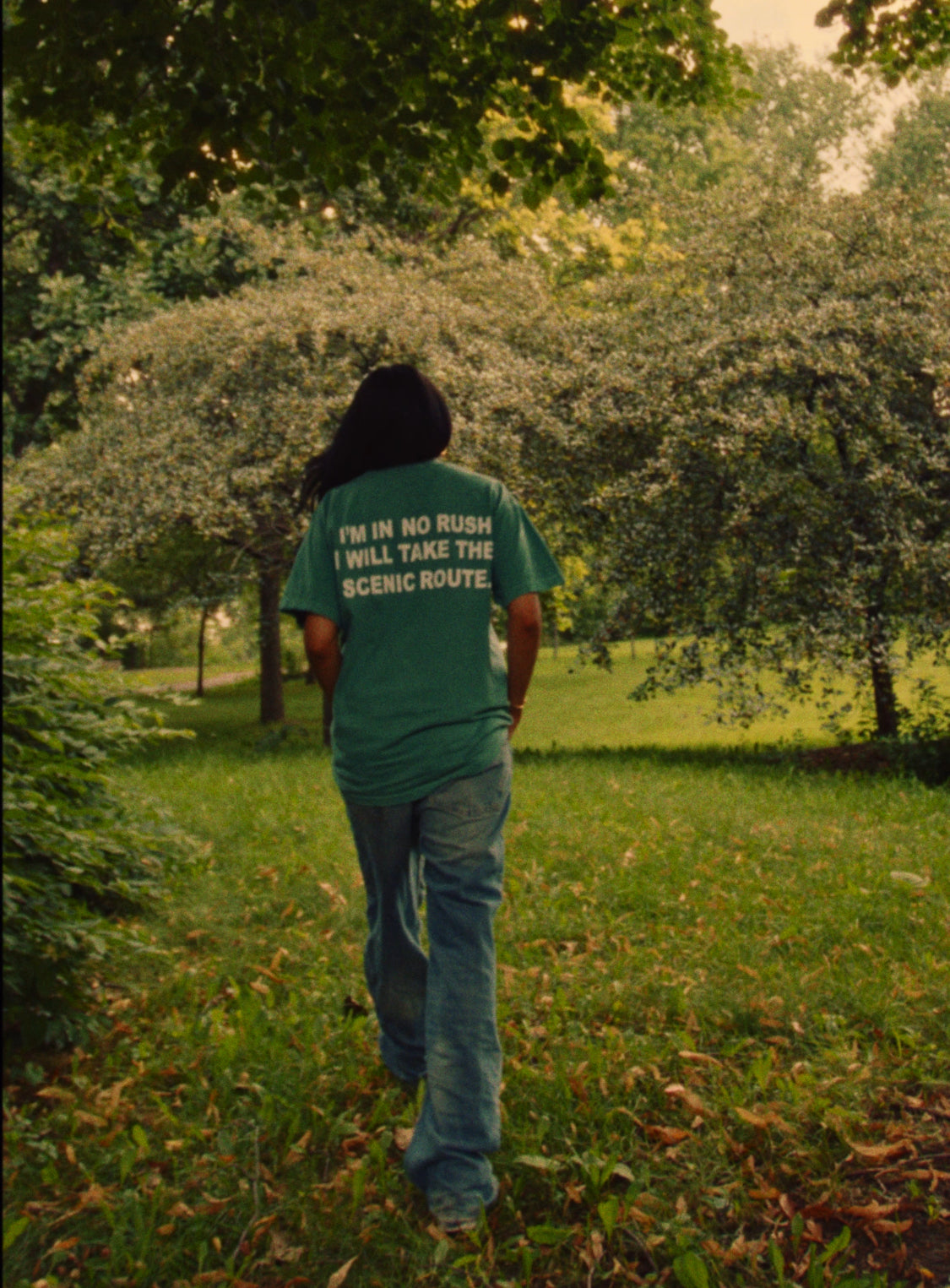 Someone walking away through a park under blossoming trees, wearing the green Entry 001 tee with jeans.